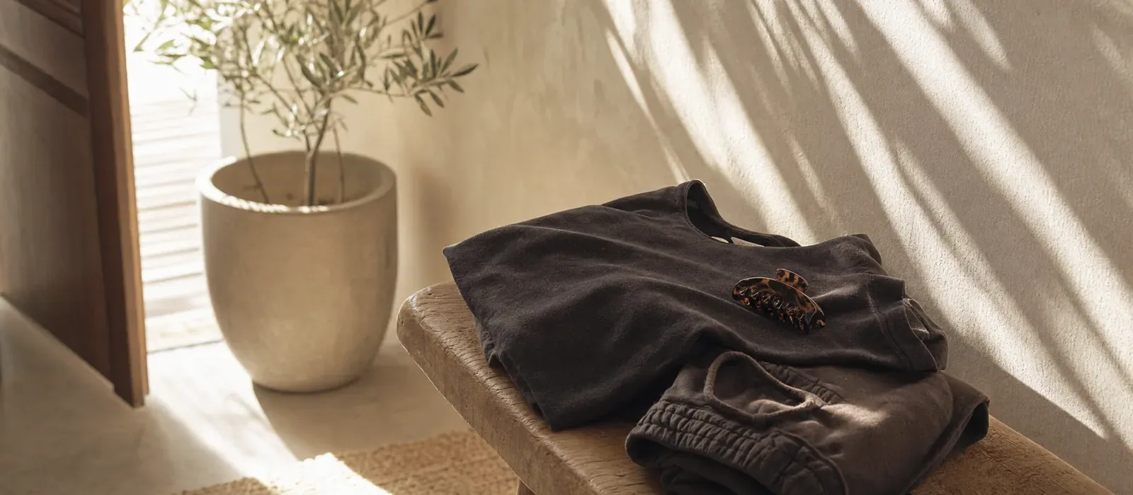 A folded dark tee and trackpants with a tortoiseshell claw clip on a timber bench in warm afternoon light by an open front door
