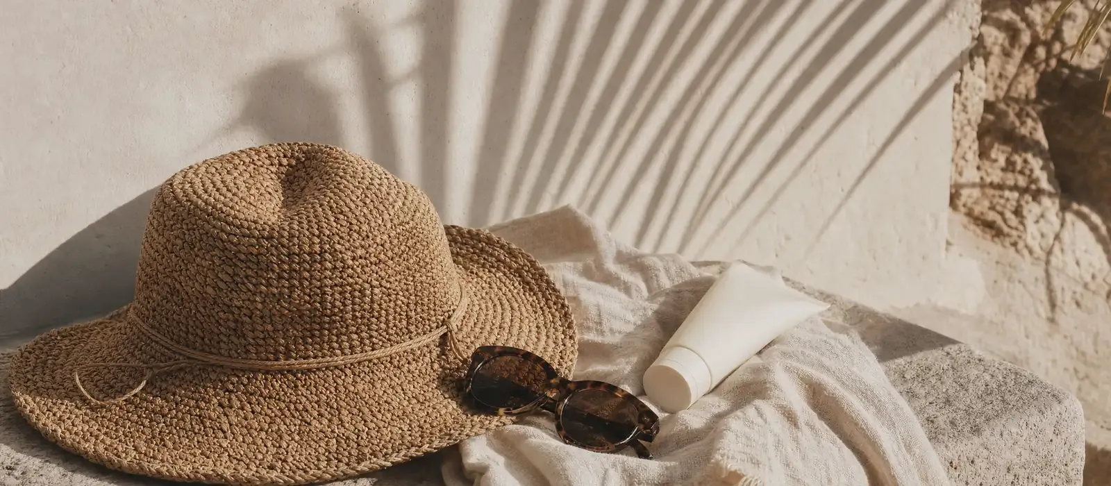 Straw sun hat, tortoiseshell sunglasses and sunscreen resting on a linen towel, palm shadows on the wall behind